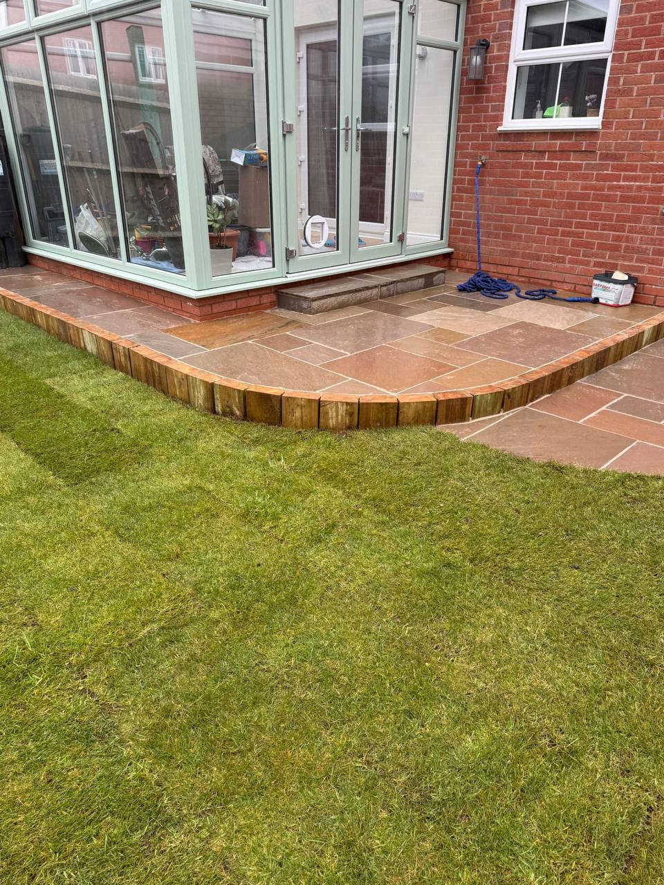 Curved raised Indian sandstone patio with timber sleeper edging next to a conservatory and fresh lawn